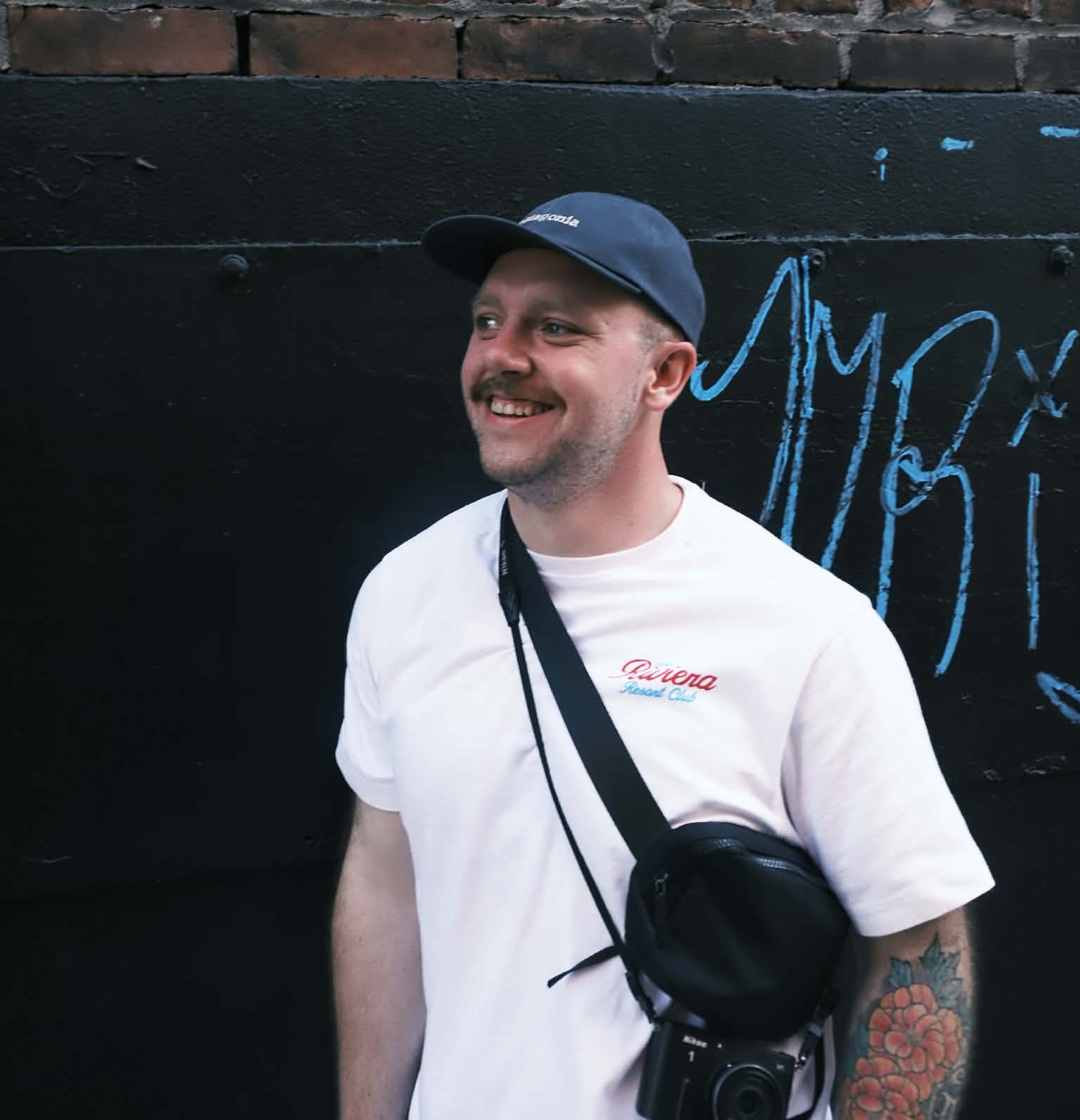 Photo of a man in front of a black and brick wall. There's graffiti on the right side. The man is wearing a white shirt and blue hat.